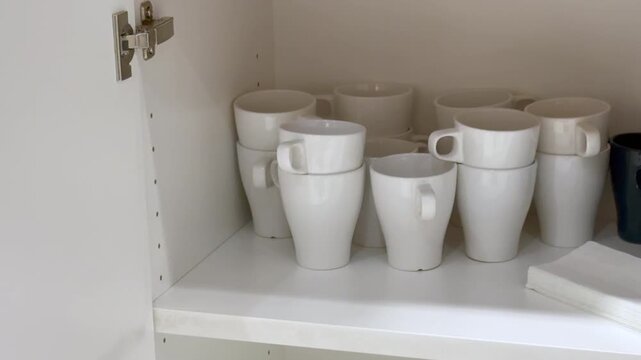 Camera moves left to right revealing white ceramic mugs stacked in an office kitchen cabinet