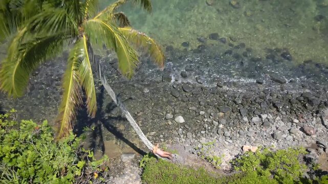 Aerial view of woman sunbathing on palm tree over tropical rocky beach shoreline.