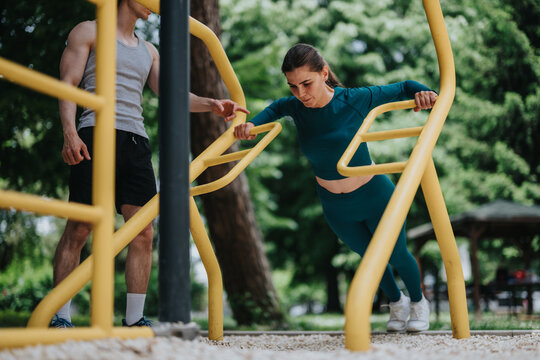 Woman performs inclined push-up on parallel bars while a trainer coaches her technique. The pair work together in an outdoor fitness park for strength training and form practice.