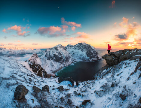 Hiker Standing on Mount Ryten Snowy Mountain Peak Overlooking Fjord at Sunset Lofoten Islands Norway