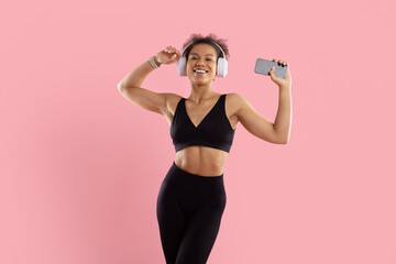 Obraz premium Happy young Black woman in fitness wear dancing with wireless headphones and smartphone, enjoying music against bright pink studio background with generous copy space