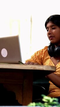 Indian Asian teenage girl studying online at home using a laptop computer while sitting by a bright, airy window in a cozy lavish home, focused on education, exam preparation, learning with dedication