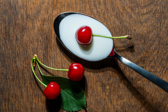Ripe red cherry on metal spoon filled with white yogurt or cream, closeup. Fresh berries with green leaf on rustic wooden table. Perfect for dairy product advertising and healthy diet concepts