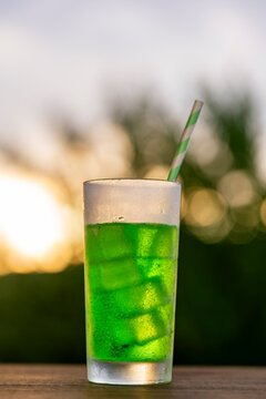 Chilled green soda drink with ice in frosted glass against beautiful sunset bokeh background, closeup. Refreshing summer cocktail with striped straw. Perfect for beverage advertising and bar menus