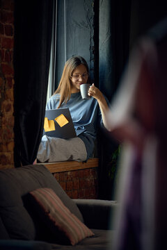 Young woman enjoying coffee while working remotely from cozy window workspace. Concept of peaceful morning routine, remote work, digital lifestyle and work life balance.