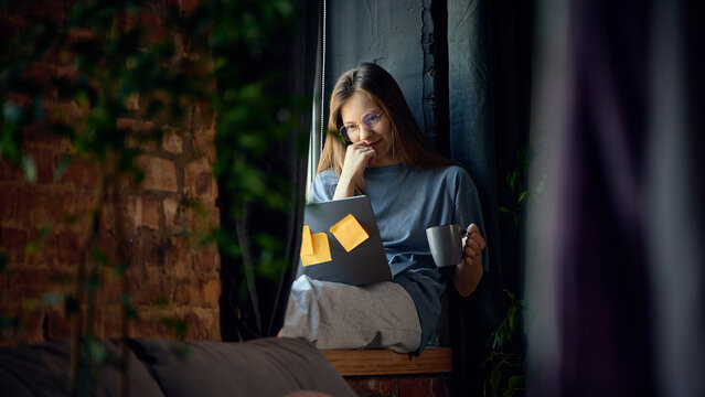 Thoughtful young woman working on laptop with coffee beside loft apartment window. Concept of online customer support, virtual consultations, digital administration and home based business.