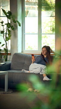 Young woman relaxing on sofa while browsing smartphone near large window. Concept of mobile shopping, product discovery and personalized retail recommendations.