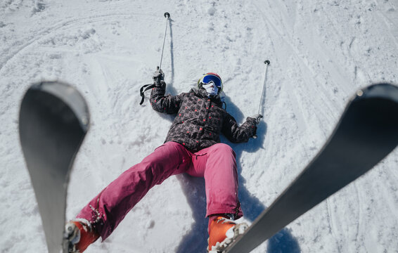A skier lies on a snow-covered slope wearing helmet, goggles and pink ski pants. The fallen skier rests on their back with skis and poles visible on the snow.