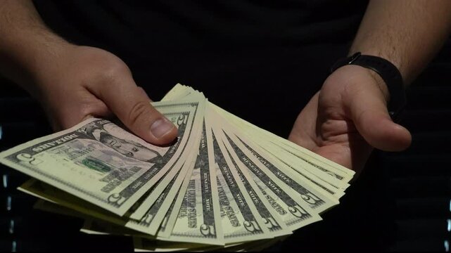 A man counts a fan of five dollar bills in his hands against a dark background. Cinematic lighting highlights the currency, suitable for finance, startup, and wealth concepts.