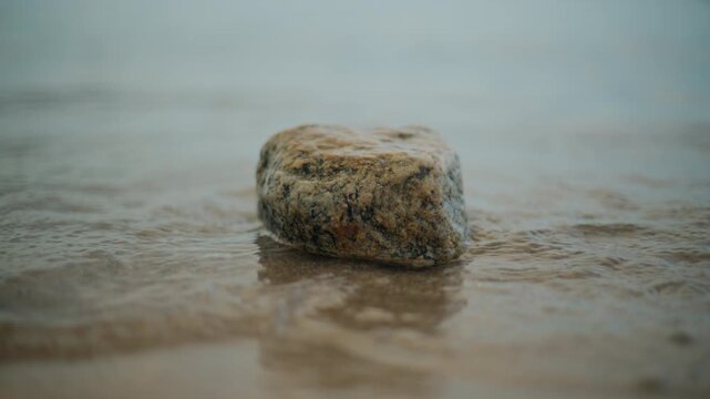 Solitary rock resisting water waves flowing over a sandy shore, symbolizing strength, resilience, and calm nature