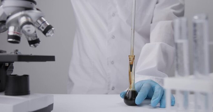 Slow motion gloved hand pipetting dark oil liquid into glass flask on bench with microscope in chemical laboratory.