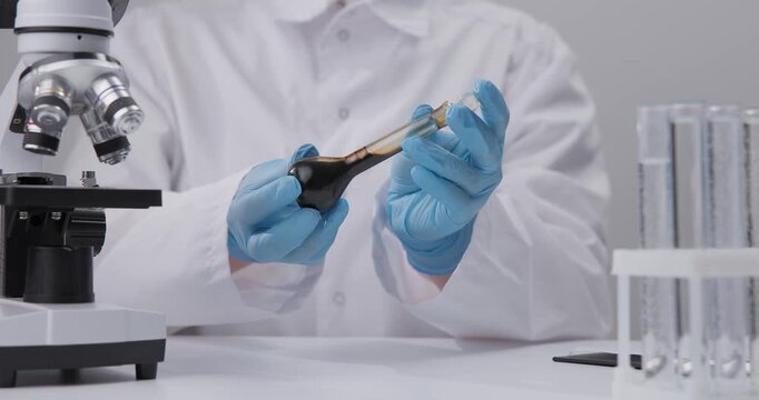 Scientist handling dark liquid oil with dropper on chemical laboratory bench near microscope and test tubes.