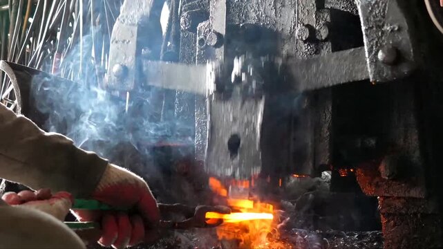 The blacksmith employs a powerful mechanical hammer to shape glowing metal on an anvil, followed by intense reheating within a coal fire furnace for further industrial forging operations.