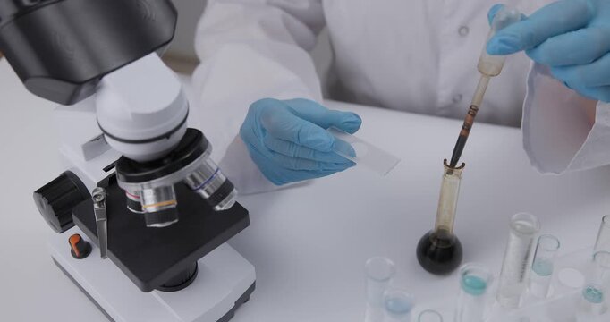 Gloved hands applying oil liquid sample onto glass slide beside microscope on laboratory bench..