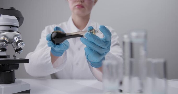 Scientist manipulating oil dark liquid in glass vial on laboratory benchtop with microscope in chemical laboratory.
