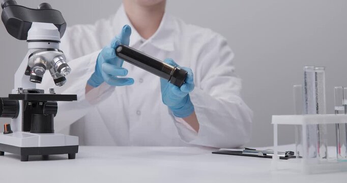 Static shot scientist handling oil sample vial at laboratory bench with microscope gloved hands rotating tube in chemical test laboratory.