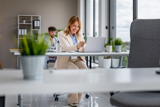 Businesswoman in modern office smiling at laptop during coworking meeting with colleagues