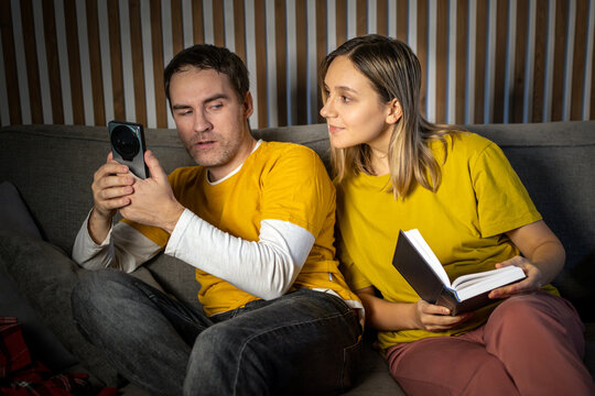 Woman hiding behind a book, secretly peeking at her partner's smartphone screen. Suspicion, jealousy, relationship control, and privacy invasion.