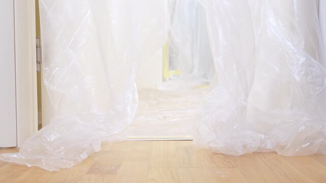 A black and tan dachshund dog in an orange construction helmet and vest pushes through hanging plastic sheeting on a wooden floor, a concept of home renovation.
