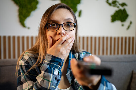 Young woman covering her mouth in shock while holding a remote control, watching something surprising on TV. Shocking movie scene, unexpected plot twist, or scary news.