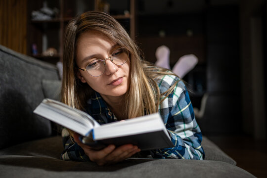 Young woman reading a book while lying comfortably on a sofa at night. Leisure reading, home hobby, and quiet evening relaxation.