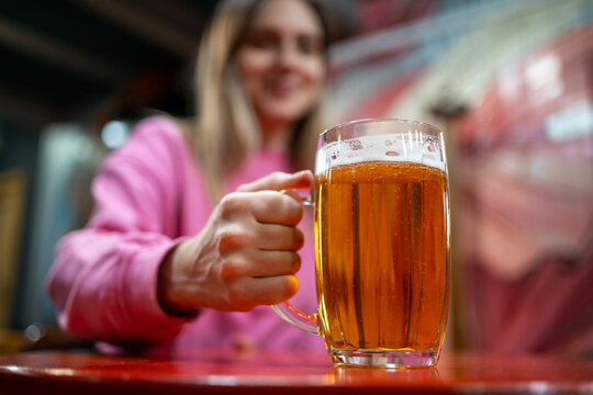 Young woman holding a glass mug of light beer in a pub or bar. Close-up of a refreshing alcoholic drink with selective focus, social leisure, nightlife, relaxation and beverage concept.