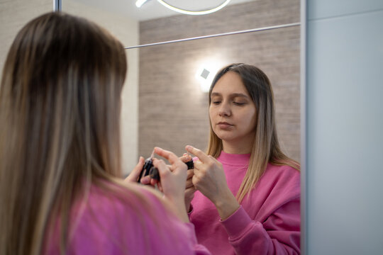Woman looking at a cosmetic product in her hands while standing in front of bathroom mirror. Beauty routine, self-care, and morning skincare prep.