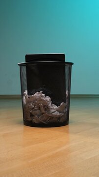 Vertical video black wire mesh trash bin standing on a wooden floor, partially filled with crumpled white papers and documents, symbolizing waste, failure, discarded ideas, and office clean up