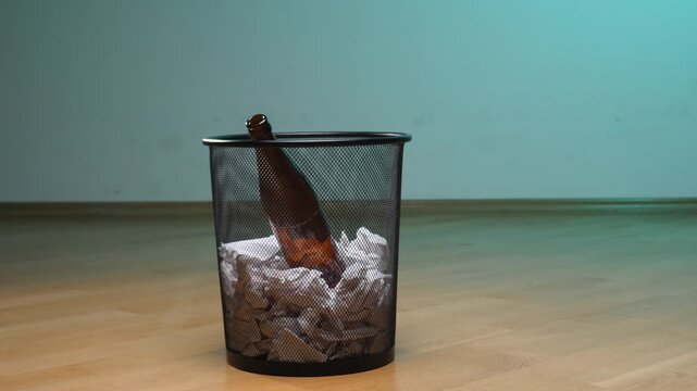 Person is throwing an empty glass bottle into a metal waste bin filled with crumpled paper, symbolizing giving up alcohol, breaking an addiction, or recovering from a bad habit