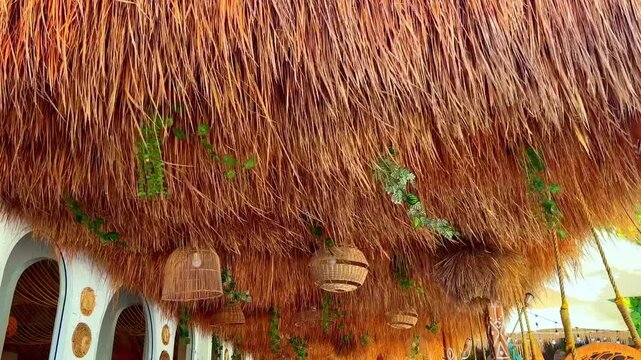 Beach restaurant interior with straw thatch roof and wicker lamps. Low angle view of a cozy outdoors cafe ceiling. Tropical resort terrace design. Exotic boho summer vacation concept.