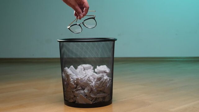 Person's hand dropping prescription eyeglasses into a metal wastebasket filled with crumpled paper, symbolizing freedom from vision correction, successful eye surgery, or getting rid of old habits