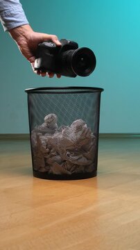 Vertical video hand discarding an obsolete digital single lens reflex camera with a lens into a black metal mesh trash bin filled with crumpled paper, symbolizing technological obsolescence and waste