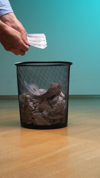 Vertical video hands dropping dollar banknotes into a waste basket filled with crumpled paper, symbolizing financial loss, reckless spending, and the devaluation of currency