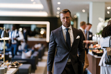Fototapeta premium A strong-looking man with a determined expression, dressed in a sleek business suit, walking confidently through a networking event filled with vibrant interactions.