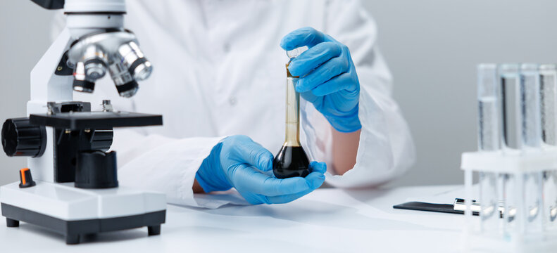 Laboratory workspace with microscope and gloved hands holding a volumetric flask of dark oil for scientific testing and analysis