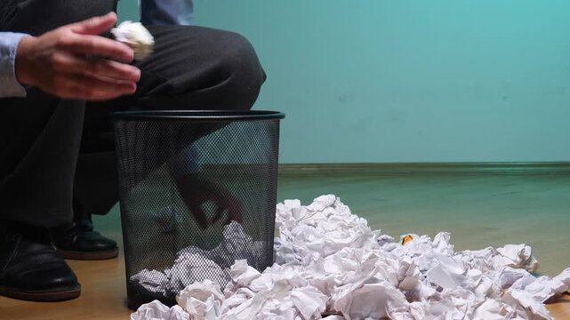 Person's hands dropping crumpled paper into a metal trash can, surrounded by more discarded paper balls on a wooden floor, representing frustration, failure, and wasted effort