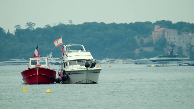 Boats anchored in a serene bay, featuring the national flags of lebanon and france, with a coastline and a historic building in the background