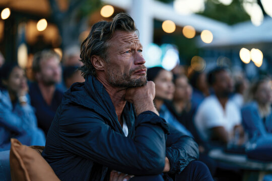 A contemplative man appears lost in thought while attending an outdoor social event, surrounded by an atmospheric backdrop filled with warm lights and engaging company.