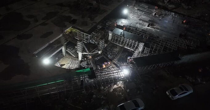 Night shot of a construction site for a new real estate complex, illuminated by bright floodlights, with workers visible among the steel framework and scaffolding.