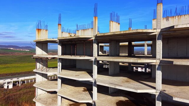 Abandoned concrete building skeleton with exposed rebar, Pomorie, Bulgaria. Aerial drone view shows rusted columns, open floors and green fields under blue sky.