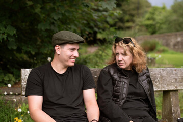 Fototapeta premium A middle-aged woman and young man her son sit on a weathered park bench in a green countryside setting, sharing a thoughtful, serious conversation, they are talking and laughing