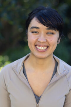 African American woman smiling at camera in garden, wearing beige jacket with zipper, stud earrings