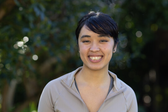 African American woman smiling, standing in park, wearing beige zip jacket, gray top, small studs