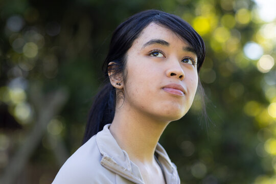 Adult female looking upward in park, holding head up, wearing light zip-up jacket and stud earring