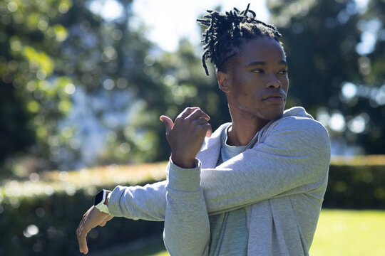 African man stretching shoulder while wearing light gray hoodie and smartwatch in sunlit park