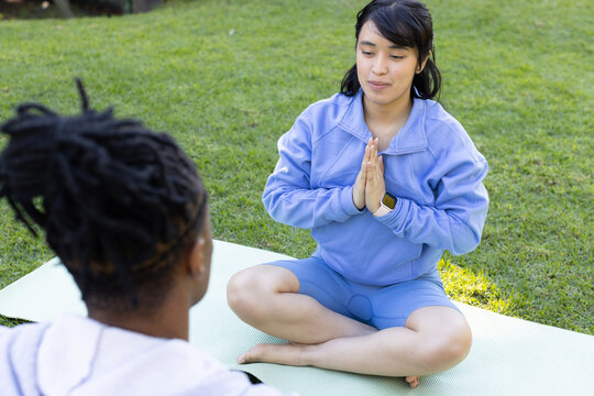 African American friends sitting in workout set doing namaste on pale green mat in park, smartwatch