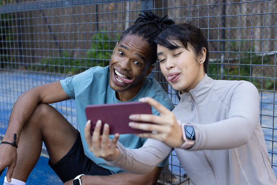 African American man and Asian woman sitting on blue court holding maroon phone and showing watch