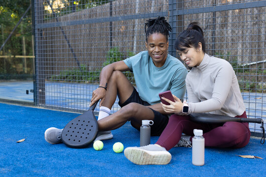 Diverse athletic friends sitting on blue sport court checking smartphone with paddles and balls