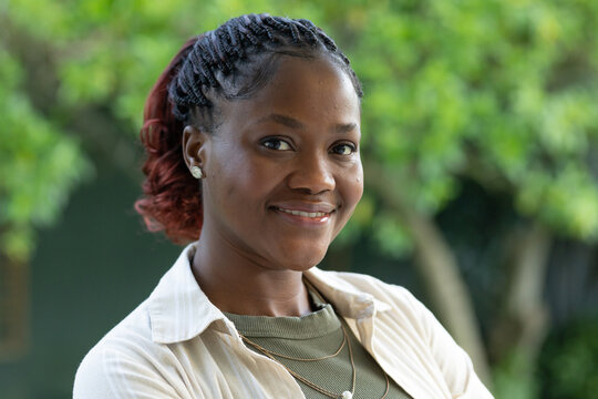 African woman smiling, posing outdoors in garden wearing beige collared shirt and gold necklaces