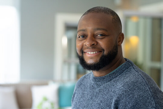 Mature African man standing facing camera in lounge, smiling, wearing grey sweater near sofa